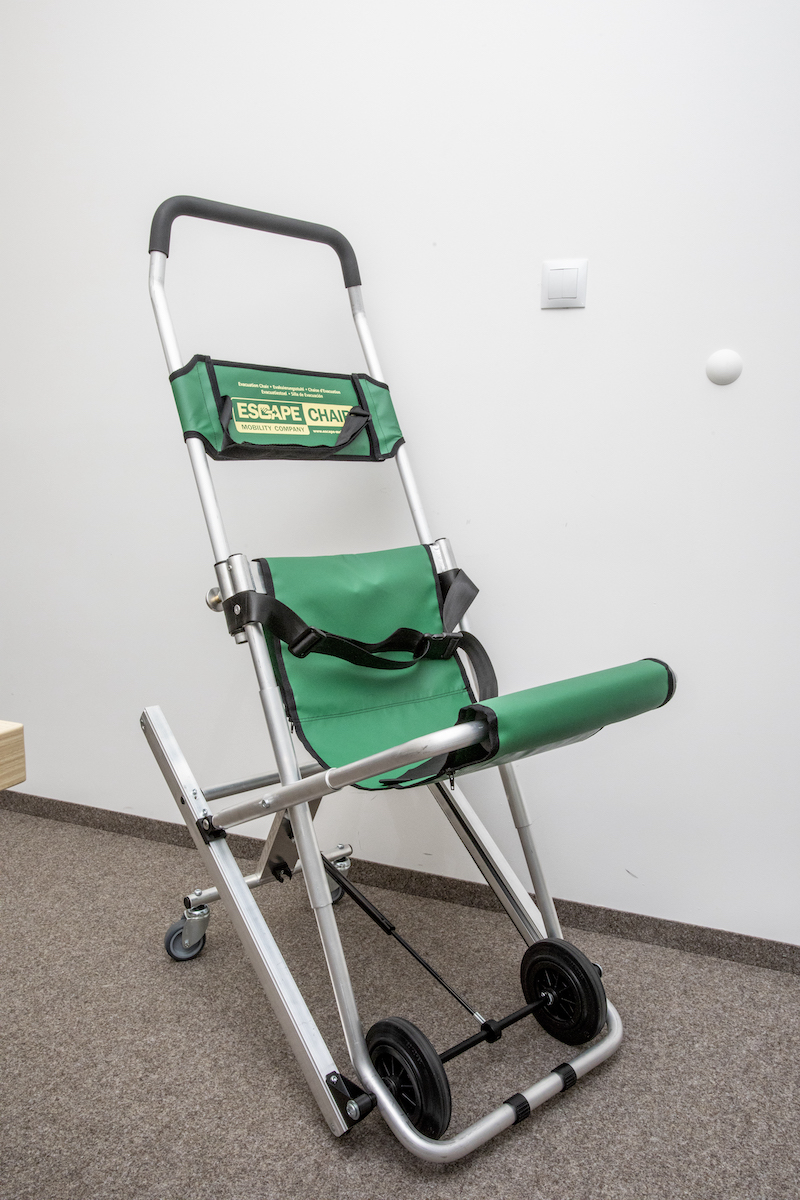 Rozłożone krzesło ewakuacyjne. Zielone materiałowe siedzenie i oparcie, metalowy stelaż z kółkami.