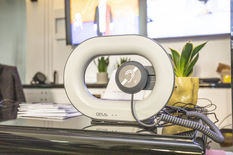 Przenośna pętla indukcyjna. Sprzęt w kształcie owalnej obręczy, pośrodku zawieszona okrągła słuchawka z symbolem pętli indukcyjnej.