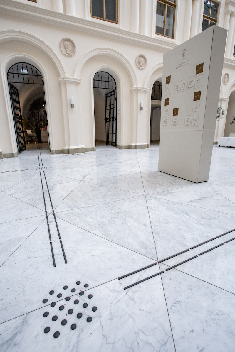 Dziedziniec. Na jasnej podłodze ciemne ścieżki dotykowe. Prowadzą do kasy. Po prawej tablica. Na niej informacja wizualna w postaci piktogramów i napisów oraz strzałki kierunkowe. 