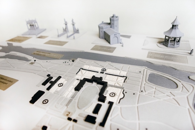 Makieta, na niej trójwymiarowe wydruki budynków i elementów ogrodu, obok mapa terenu Pałacu, wraz z ogrodami i sąsiadującymi budynkami, wszystko opisane Braillem.