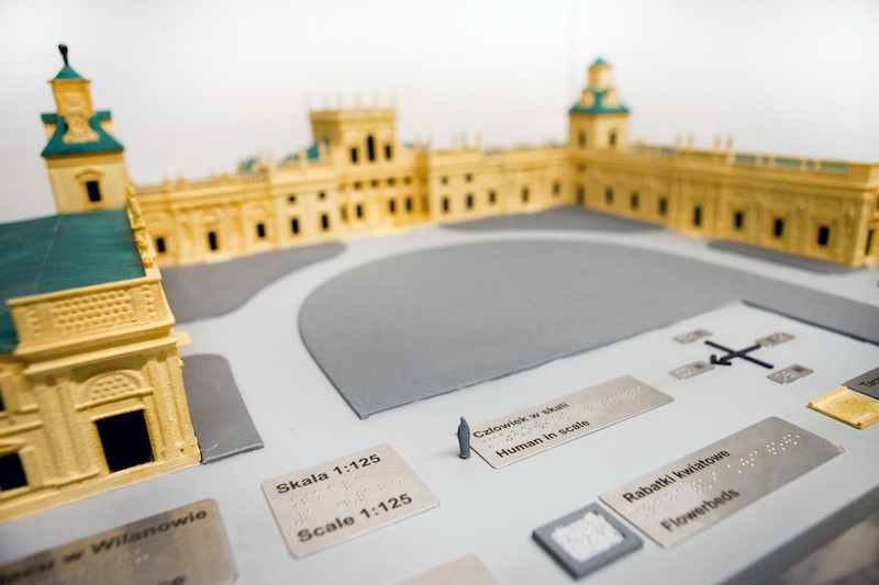Makieta Pałacu Króla Jana III w Wilanowie. Na planszy trójwymiarowy model budynku, model człowieka w skali, plakietki z opisem elementów w Braille’u.