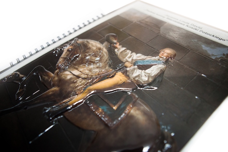 Fragment dotykowego katalogu. Plastikowa tłoczona tyflografika, przedstawia mężczyznę na koniu, nad nią napis w czarnodruku i w Braille’u.