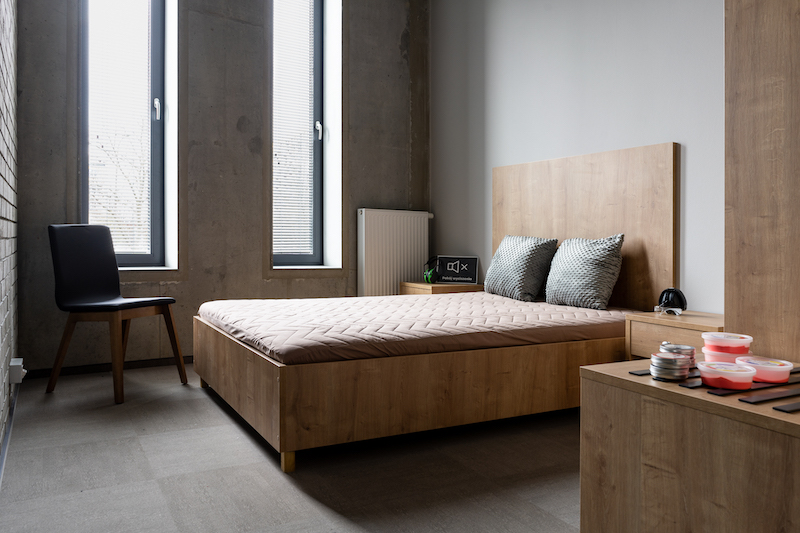 Wnętrze pokoju wyciszenia, przypomina pokój hotelowy. Pośrodku pokoju duże łóżko z dwiema poduszkami, obok krzesło i szafki.