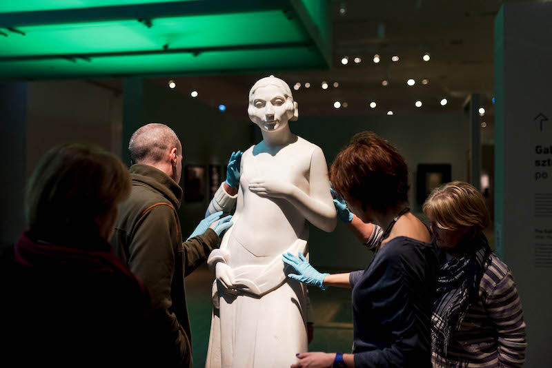Wysoka rzeźba przedstawiająca postać. Dookoła kilka osób w lateksowych rękawiczkach, dotykają rzeźby z różnych stron.