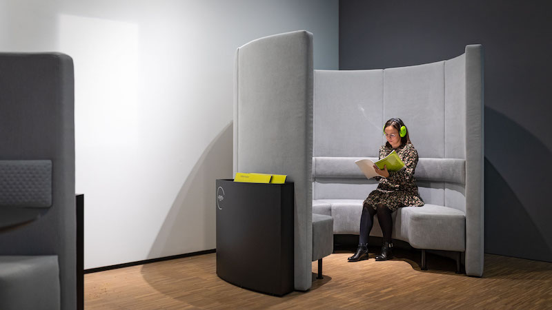 W rogu sali ustawiony półokrągły segment składający się z kanapy i wysokich miękkich ścianek, będących częścią oparcia kanapy. W środku siedzi osoba, ma na głowie słuchawki wyciszające, czyta folder.