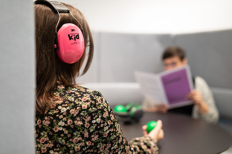 W środku półokrągłego segmentu składającego się z kanapy i wysokiego oparcia, tworzącego ściany, siedzą dwie osoby. Jedna ma na głowie słuchawki wyciszające, w ręku trzyma miękką piłeczkę sensoryczną, druga osoba czyta folder.