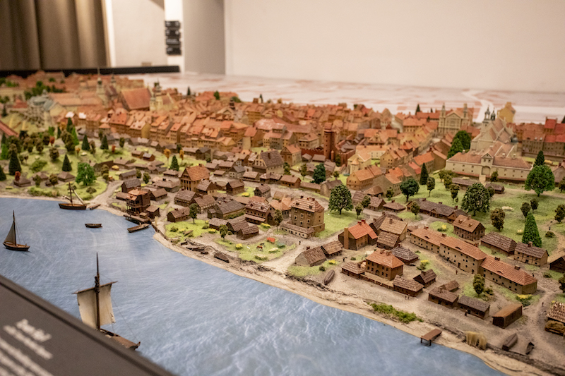 Muzeum Warszawy. Zbliżenie na makietę. Przy brzegu rzeki szeregi drewnianych domów, drewnianie pomosty, na rzece łodzie żaglowe.