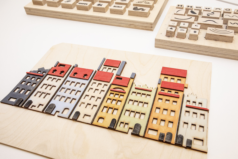 Drewniana plansza z wypukłymi kolorowymi kształtami elewacji kamienic Starego Miasta. Zaznaczone są drzwi, okna, dachy.