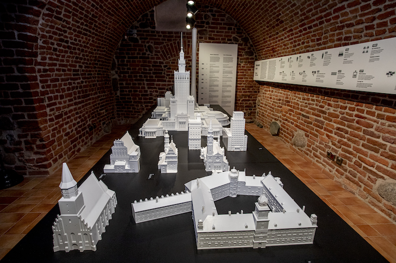 Muzeum Warszawy. Na długim czarnym stole ustawione kilkanaście wydruków 3D najważniejszych budynków Warszawy. Wśród nich Pałac Kultury i Nauki, Pałac Królewski, kilka kościołów.