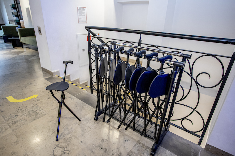 Balustrada przy schodach. Niewielki stojak, na nim ustawione przenośne rozkładane taborety. Złożony taboret może działać jak laska do wspierania się, na rozłożonym można przysiąść.