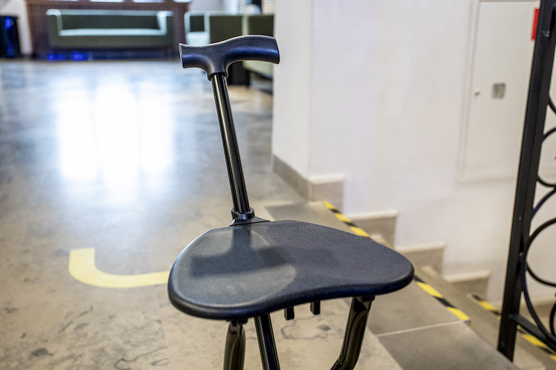 Rozstawiony taboret. Ma trzy nogi, niewielkie siedzenie. Jedna z nóg jest przedłużona w oparcie, zakończona rączką ułatwiającą chwycenie. Przypomina laskę do wspierania się.