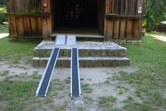 Wejście do zabytkowego drewnianego budynku. Do środka prowadzi kilka kamiennych stopni i drewniany próg. O próg oparte są dwie wąskie przenośne szyny.