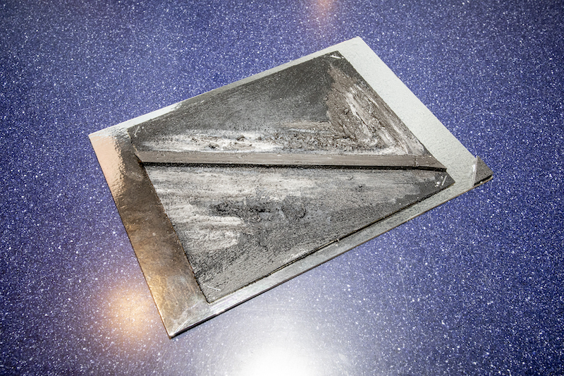 Tyflografika obrazu abstrakcyjnego. Czarne tło namalowane smugami grubej farby o wyraźnej fakturze. Pośrodku obrazu przyklejona cienka drewniana listwa.