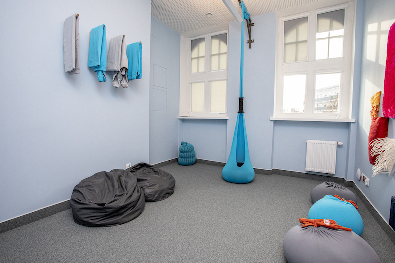 Wnętrze pokoju wyciszeń. Na podłodze szara wykładzina, ściany w kolorze błękitnym. Do sufitu przymocowane siedzisko w kształcie kokonu. Na ścianie powieszone kolorowe koce, pod ścianą ustawione czarne pufy do siedzenia.
