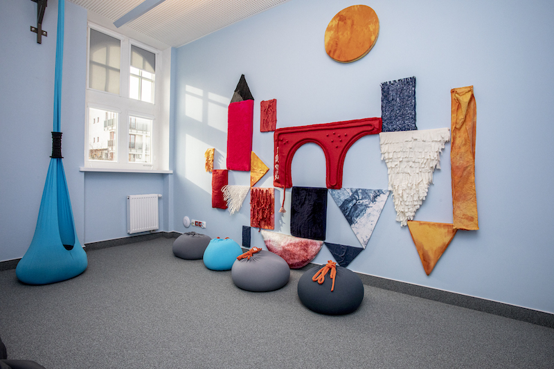 Wnętrze pokoju wyciszeń. Na podłodze szara wykładzina, ściany w kolorze błękitnym. Na ścianie dekoracja w kawałków kolorowych materiałów. Na podłodze pufy o różnych kształtach i kolorach,