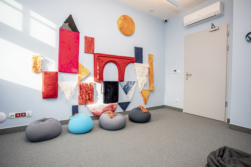 Wnętrze pokoju wyciszeń. Na podłodze szara wykładzina, ściany w kolorze błękitnym. Na jednej ze ścian dekoracja z kawałków materiału w kształcie zamku z wieżami. Pod ścianą ustawione pufy do siedzenia.