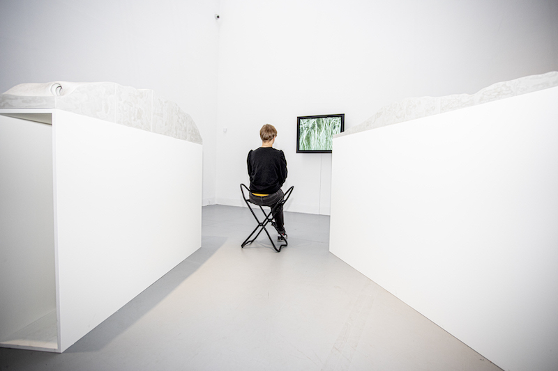 Przestrzeń wystawy. Przed eksponatem osoba, siedzi na składanym taborecie.