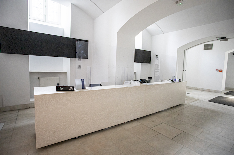 Korytarz przy wejściu do Zachęty. Długa biała lada, za nią rząd ekranów. Na ladzie tabliczka z napisem „Info” i symbolem pętli indukcyjnej.
